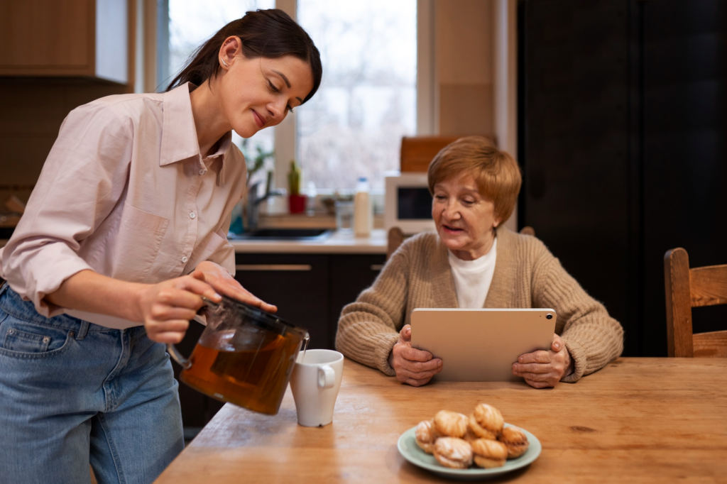 A caregiver pouring a cup of some drink for an elderly person sitting at the table and using a tablet