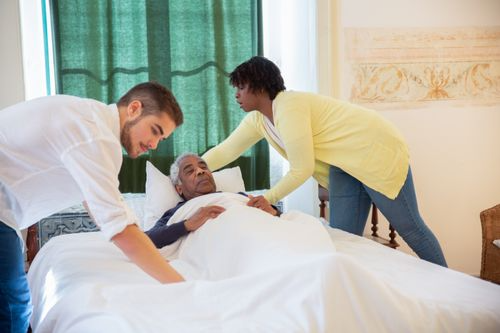 Two caregivers helping an old man get into bed